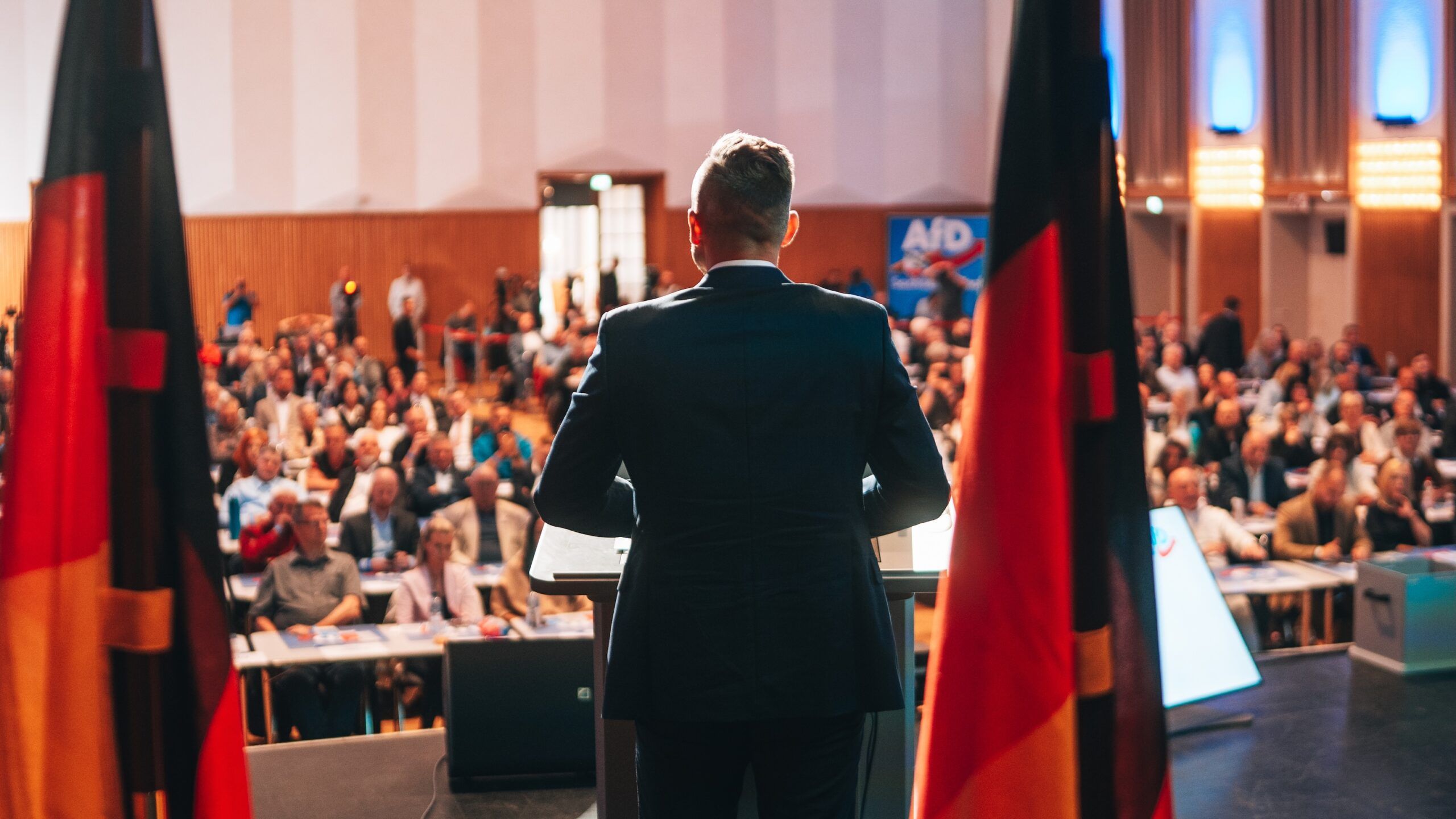 Ulrich Siegmund steht mit dem Rücken zur Kamera an einem Rednerpult vor einem voll besetzten Saal